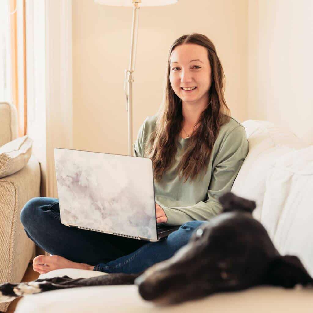 Estelle sitting cross-legged on a white sofa, with a laptop on her lap. The face of her greyhound Buster is visible in the foreground but is out of focus.