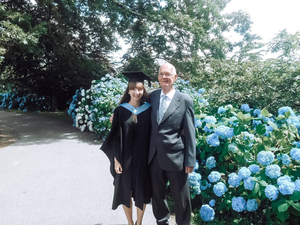 Estelle in a black graduation gown with a blue collar, standing next to her dad and in front of a blue hydrangea bush