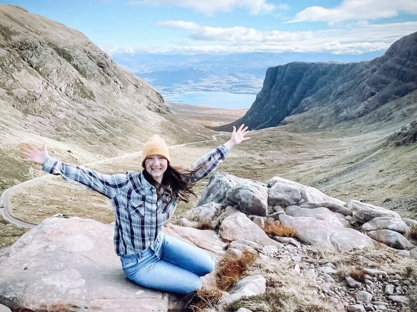 Estelle sat on a rock with her arms outstretched, with a winding road and mountains and a loch behind her